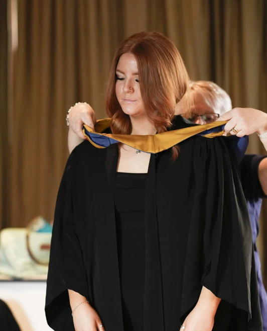 A staff member placing a gold stole over a female graduate's gown during the graduation preparations. A staff member placing a gold stole over a female graduate's gown during the graduation preparations.
